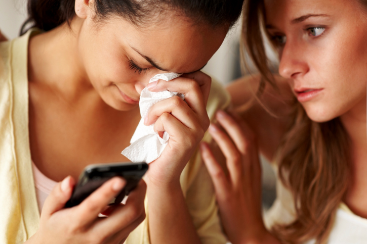 A woman holding a tissue to her face, crying, and holding her cellphone. Another woman is comforting her.