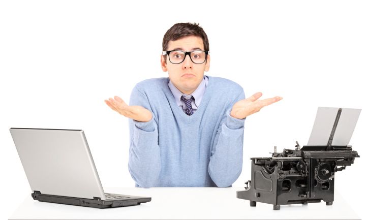 a man in glasses, a button down shirt, tie, and a pullover sweater with his arms bent at the elbows as if to say, 'I don't know.' On one side of him is a laptop computer and one the other side is a typewriter.