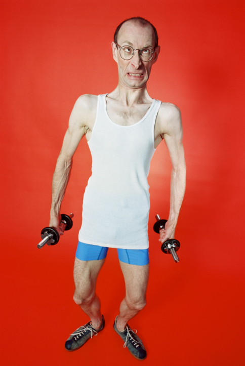 Very tall, very thin man with glasses and a pained expression wearing a white tank top, small blue shorts, and sneakers and holding a set of very small, light hand weights. 