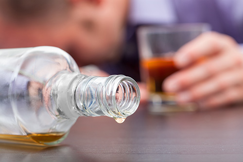 An almost empty bottle of alcohol turned on its side. A droplet of alcohol is hanging off the rim. In the background, a man with a glass of alcohol is resting his head on the table.